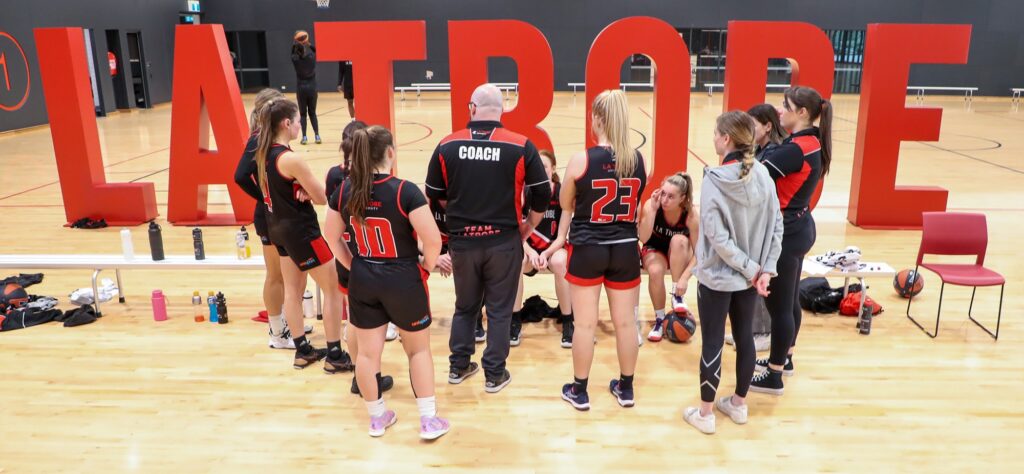 Basketball coach addressing players in a huddle on an indoor court with large red letters in the background.